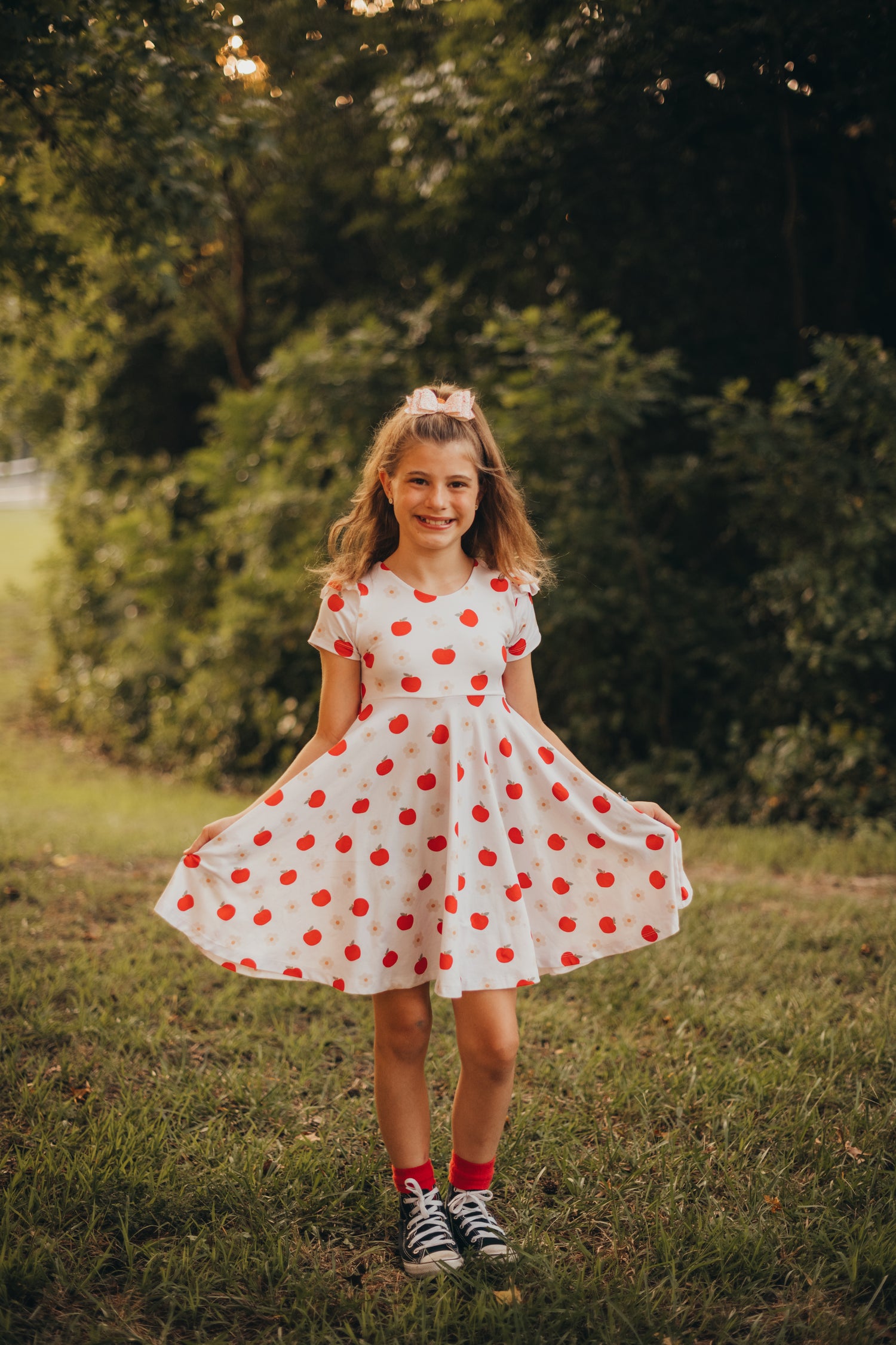 Girl wearing dress with apples on it, converse sneakers and red socks, wearing a hair bow smiling